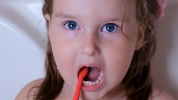 Ένα κοριτσάκι 3 ετών βουρτσίζει τα δόντια της με μια κόκκινη οδοντόβουρτσα στο μπάνιο του σπιτιού. Υγειονομική περίθαλψη, οδοντιατρική υγιεινή, άνθρωποι και ομορφιά — Φωτογραφία Αρχείου