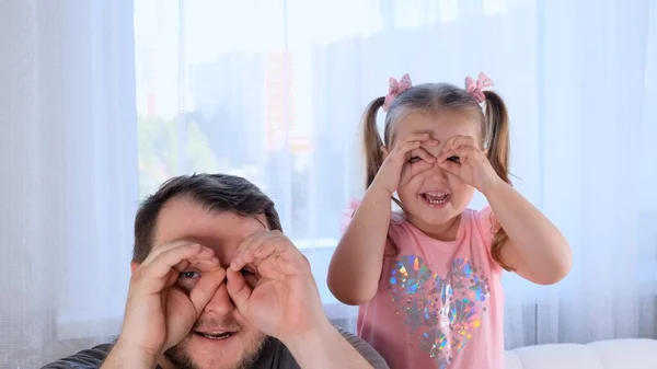 Lustige Kleine Tochter 3 Jahre alt und Vater haben Spaß zu Hause, machen lustige Gesichter, basteln Brillen mit ihren Fingern, wie Brillen, die durch Ferngläser in die Kamera schauen. Viel Spaß mit Kindern — Stockfoto