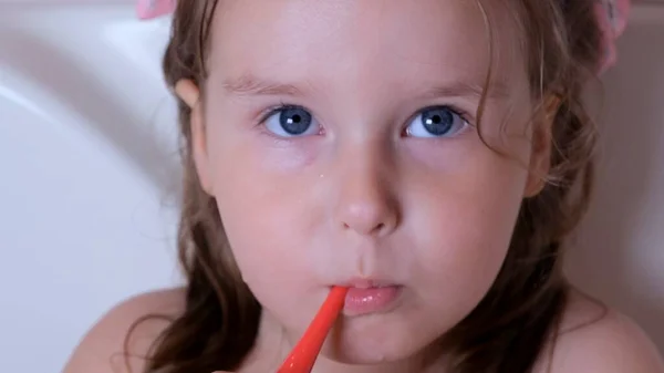 Ένα κοριτσάκι 3 ετών βουρτσίζει τα δόντια της με μια κόκκινη οδοντόβουρτσα στο μπάνιο του σπιτιού. Υγειονομική περίθαλψη, οδοντιατρική υγιεινή, άνθρωποι και ομορφιά — Φωτογραφία Αρχείου