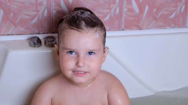 Das kleine lachende Mädchen, 3 Jahre alt, badet und wäscht sich im heimischen Badezimmer mit Schaum auf dem Kopf im Wasser. Babypflegekonzept, Hygiene. — Stockfoto