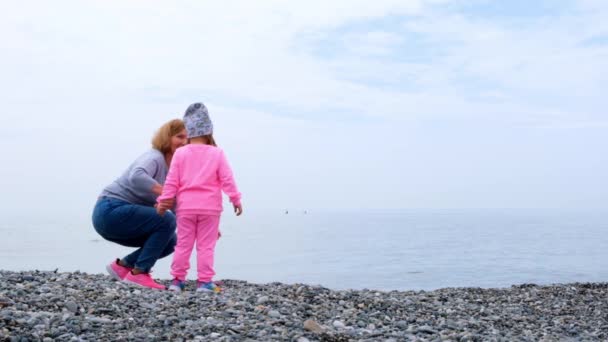 Nonna e nipote camminano sulla riva del mare, giocano, lanciano sassi e si divertono in una giornata autunnale di primavera. Il concetto di infanzia, genitorialità, relazioni tra figli e genitori — Video Stock