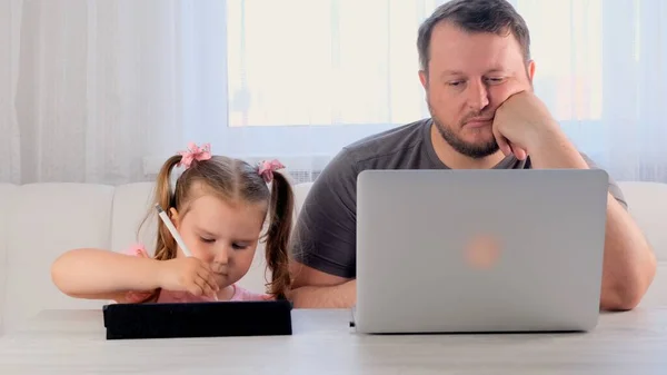 Uttråkad far till en affärsman arbetar på en bärbar dator hemma bredvid en liten flicka, 3 år gammal, spelar i tabletten. Modern familj. Frilansbegreppet. Beroende av prylar — Stockfoto