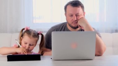 Sıkılmış bir iş adamının babası evde dizüstü bilgisayarda çalışıyor. Yanında 3 yaşında bir kız var. Tablette oynuyor. Modern aile. Serbest meslek kavramı. Aygıtlara bağımlılık