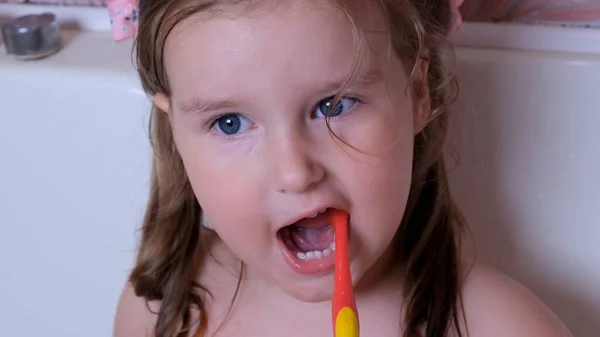 Niña divertida de 3 años se cepilla los dientes con un cepillo de dientes rojo en el baño en casa. Salud, higiene dental, personas y concepto de belleza —  Fotos de Stock