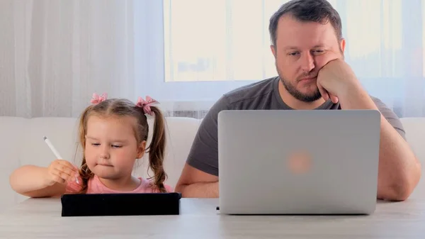 funny father of a businessman is working on a laptop at home next to a Little girl, 3 years old, playing in the tablet. Modern family. Freelance concept. Addiction to gadgets