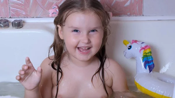 Kleines lachendes Mädchen, 3 Jahre alt, mit zwei Pferdeschwänzen auf dem Kopf, badet und wäscht zu Hause im Badezimmer im Wasser. Babypflegekonzept, Hygiene. — Stockfoto