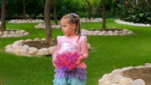 En liten jente på tre år, med to hestehaler på hodet, kledd i en skjør, rosa blå kjole, leker med en lys, gjennomsiktig ball med flerfargede fjær inni. – stockvideo