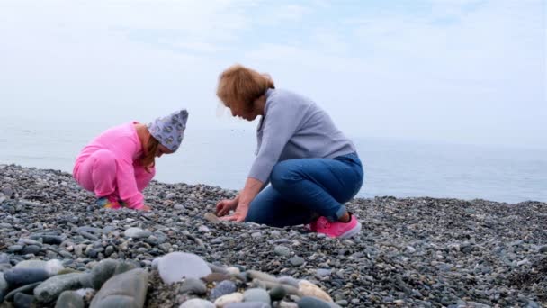 Nonna e nipote camminano sulla riva del mare, giocano, lanciano sassi e si divertono in una giornata autunnale di primavera. Il concetto di infanzia, genitorialità, relazioni tra figli e genitori — Video Stock