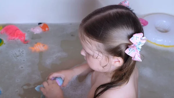 Kleines spielendes Mädchen, 3 Jahre alt, mit zwei Pferdeschwänzen auf dem Kopf, badet und wäscht sich im heimischen Badezimmer im Wasser. Babypflegekonzept, Hygiene. — Stockfoto