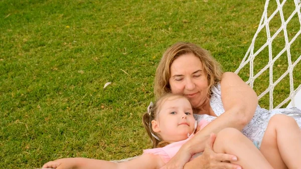 Das Ehepaar Großmutter und Enkelin 3 Jahre alt relaxen und sonnen sich an einem sonnigen Sommertag in einer Hängematte auf einer grünen Wiese in ihrem Garten. Urlaubskonzept, Generationenbeziehung, Elternschaft — Stockfoto