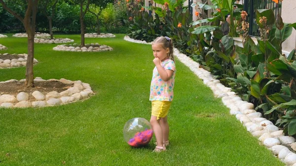 Sød lille pige, 3 år gammel, med to hestehaler på hovedet, iført en multi-farvet T-shirt og gule shorts, leger med en multi-farvet bold på en grøn eng i haven af hendes hus. Den - Stock-foto