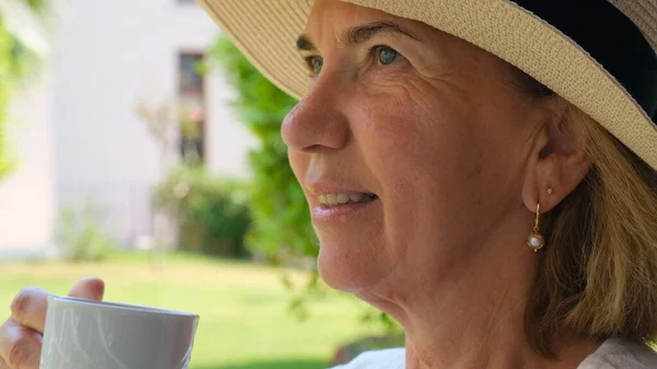 Femme adulte âgée de 50 à 55 ans dans un chapeau de paille boit une boisson chaude de café ou de thé dans une tasse blanche tôt le matin sur la terrasse de sa maison dans un jardin verdoyant par une journée ensoleillée, gros plan. Senior senior — Photo