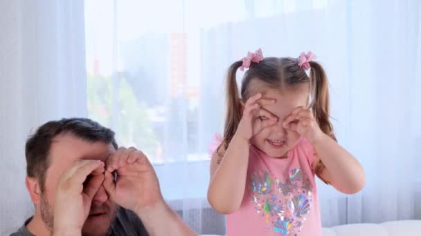 Смешная маленькая дочь 3 года и отец веселятся дома, делают смешные лица, делают очки пальцами, как очки, смотрят в камеру через бинокль, смотрят друг на друга — стоковое видео