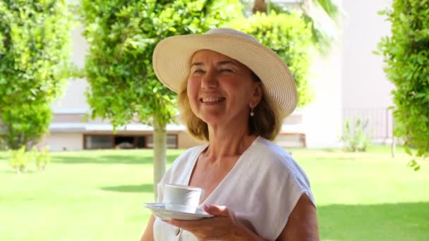 Dromer Een volwassen vrouw van 50-55 jaar in een strohoed drinkt 's morgens vroeg op het terras van haar huis in een groene tuin op een zonnige dag een warme borrel koffie of thee uit een wit kopje. Senior senior — Stockvideo