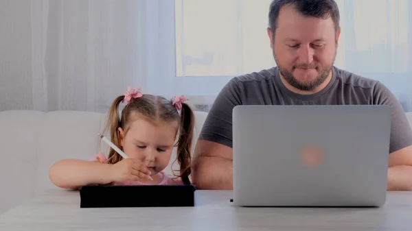 Pai triste de um homem de negócios está trabalhando em um laptop em casa ao lado de uma menina, 3 anos, jogando no tablet. Família moderna. Conceito freelance. Vício em gadgets — Fotografia de Stock