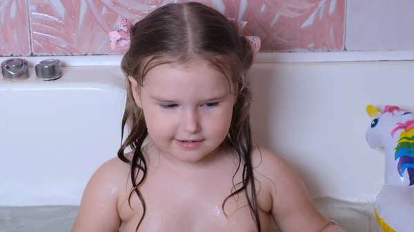 Kleines spielendes Mädchen, 3 Jahre alt, mit zwei Pferdeschwänzen auf dem Kopf, badet und wäscht sich im heimischen Badezimmer im Wasser. Babypflegekonzept, Hygiene. — Stockfoto