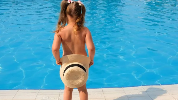 Kleines Mädchen steht und bedeckt sich mit einem Sonnenhut vor dem Hintergrund des blauen Wassers des Pools oder Meeres. Sommerurlaub, Sonnencreme, Reisen. — Stockfoto