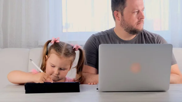 sad father of a businessman is working on a laptop at home next to a Little girl, 3 years old, playing in the tablet. Modern family. Freelance concept. Addiction to gadgets