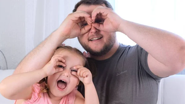 Rindo Filhinha de 3 anos e pai estão se divertindo em casa, fazendo caras engraçadas, fazendo óculos com os dedos, como óculos olhando para a câmera através de binóculos. divirta-se com criança — Fotografia de Stock