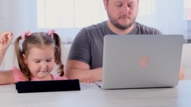 Bir iş adamının komik babası evinde, tablette oynayan 3 yaşındaki küçük bir kızın yanında dizüstü bilgisayarla çalışıyor. Modern aile. Serbest meslek kavramı. Aygıtlara bağımlılık