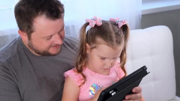 Geïnteresseerd klein meisje 3 jaar oud en haar vader studeren samen op een tablet en kijken naar een les online thuis. Papa helpt zijn dochter met haar studie. Het concept van online afstandsonderwijs. — Stockvideo