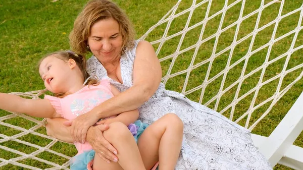 Grandmother and granddaughter, 3 years old, rest and bask in a hammock on a green meadow in their garden on a sunny summer day. Vacation concept, generational relationship, parenting, happy childhood. — Stock Photo, Image