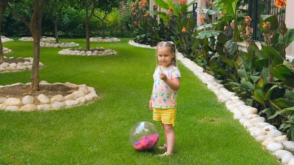 陽気かわいい女の子, 3歳,彼女の頭の上に2つのポニーテールと,マルチカラーのTシャツと黄色のショートパンツを身に着けています,彼女の庭の緑の草原でマルチカラーのボールで遊んでいます — ストック写真