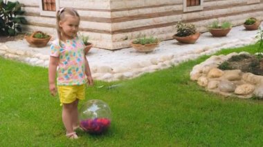 Üç yaşında, kafasında iki at kuyruğu olan, çok renkli tişört ve sarı şort giyen küçük bir kız, evinin bahçesindeki yeşil bir çayırda çok renkli bir topla oynuyor. Şey...