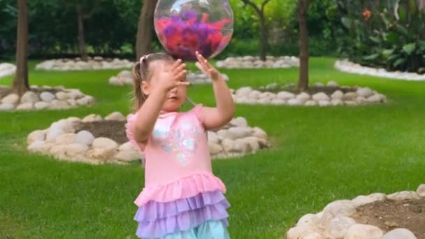 Fröhliches kleines Mädchen, 3 Jahre alt, mit zwei Pferdeschwänzen auf dem Kopf, gekleidet in ein zartes und buntes Kleid von rosa blauer Farbe, spielt mit einem leuchtend transparenten Ball mit bunten Federn — Stockvideo