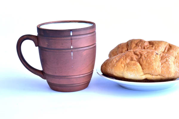 图上是一只用棕色粘土制成的杯子 牛奶倒入杯子里 牛奶滴在容器的墙壁上 旁边的盘子里有两个面包 — 图库照片