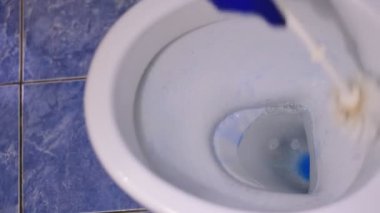 Lastik eldiven takmış bir kadın eli, beyaz bir tuvaleti fırça ve deterjanla temizliyor..