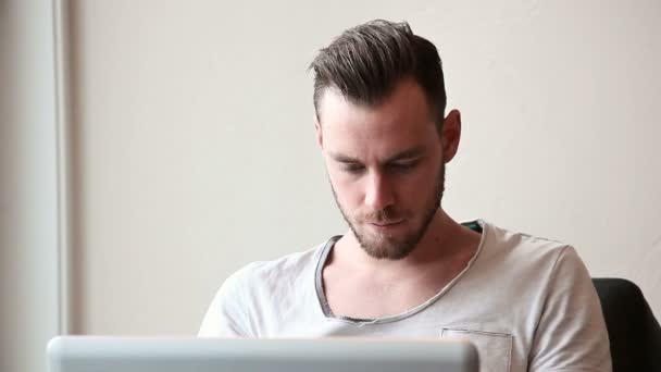 Jovem com um laptop tomando uma bebida de uma xícara de café, vestindo uma camiseta — Vídeo de Stock