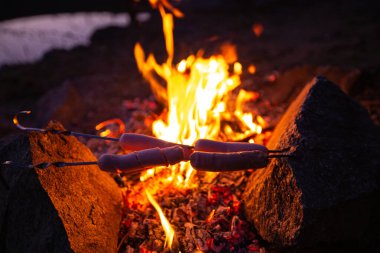 Şişte sosisler şenlik ateşinde kızarıyor ve taşlarda yatıyor. Seyahat fotoğrafçılığı. Sakin akşam