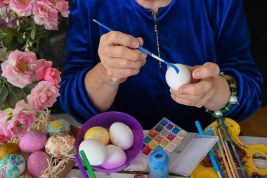 Geleneksel Paskalya yumurtası dekorasyonu için beyaz tavuk yumurtası ve mavi boyalı fırça tutan yaşlı bir kadının yakın çekimi. Fırçalar, boyalar ve süslenmiş yumurtalar masanın üzerinde duruyor..