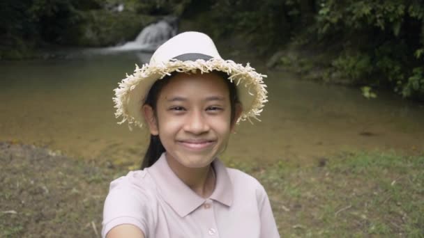 快乐的亚洲女孩 戴着草帽 微笑着 站在小瀑布边自画像 在热带雨林玩乐的漂亮女孩与朋友分享自己的视频 — 图库视频影像