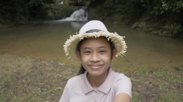 快乐的亚洲女孩 戴着草帽 站在小瀑布边自拍自画像 可爱的女孩在热带森林与朋友分享自己的视频 — 图库视频影像