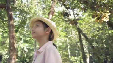 Samandan şapka takan mutlu Asyalı bir kızın etrafında dönüp güneş ışığı altında yemyeşil orman parkına bakıyor. Yaz tatili. Gezi gezisi. Tayland.