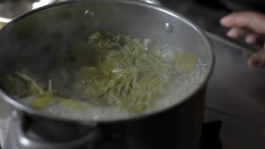 Kepçeyle kaynatılmış yerel sebzeleri fırındaki mutfak tenceresinde bir aşçı karıştırıyor. Yükselen buharla yemek pişirirken su köpürüyor..