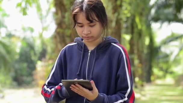公園に立っている間 青のジャケットを保持し 社会的なオンライン通信のためのデジタルタブレットを使用して健康的なタイの女の子 無線技術だ デジタルデバイスでオンラインで情報を読む — ストック動画