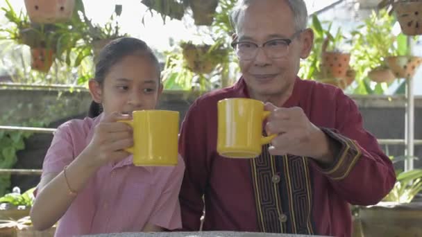 健康的な高齢者の祖父と幸せな若い愛らしい孫娘は 家の前で一緒に飲むと黄色のカップをクリックするための時間をかけています 健康的なライフスタイル — ストック動画