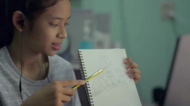 Sevimli Asyalı genç kız kulaklık takıyor ve ev bilgisayarındaki arkadaşlarına ev ödevini internetten anlatıyor. Bilgisayarlı web kameralı sohbet notlar alır. Uzaklık eğitimi kavramı.