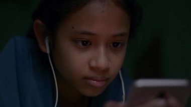 Kulaklık takan güzel Asyalı kız portresini kapat ve gece yatakta yatarken akıllı telefondan film ya da müzik videosu izlemekten zevk al. Modern yaşam tarzı. Çevrimiçi medya eğlence kavramı.