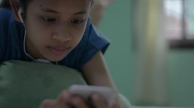 Kulaklık takan sevimli Taylandlı kız cep telefonundan sosyal medyada mesajlar ve mesajlar okumaktan zevk alıyor. Evde internet üzerinden akıllı telefon kullanırken yatakta uzanan ve dinlenen genç bir kız..