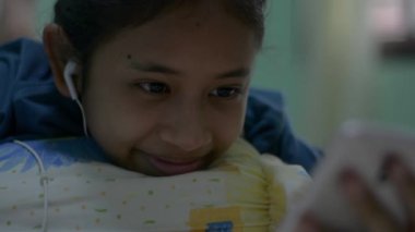 Kulaklık takan mutlu Asyalı kız ev interneti üzerinden cep telefonundan sosyal güvenlik videosu izliyor. Yatakta uzanan ve gevşeyen genç bir bayan akıllı telefondan televizyon dizisinin tadını çıkarıyor..