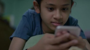 Asyalı güzel kız evdeki karantina sırasında arkadaşlarıyla sosyal iletişim üzerinden cep telefonundan mesaj atıyor. Genç kız yatakta uzanıyor ve dinleniyor. Akıllı telefon kullanmaktan zevk alıyor..