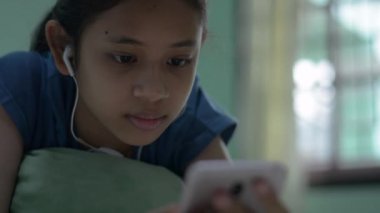 Kulaklık takan Taylandlı güzel bir kız evdeki cep telefonundan sosyal ağdan mesaj atmayı sever. Akıllı telefonlu film dizisi izlerken yatakta uzanan ve dinlenen genç bir kız..