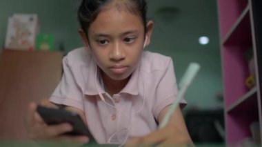 Evde karantina sırasında cep telefonundan ders almak için kulaklık takan Asyalı güzel bir kız. Dişi ergen kız yerde uzanıyor ve ödev yaparken elinde akıllı telefon tutuyor..