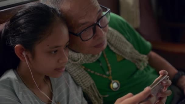 おじいさんのそばに座っているイヤホンを身に着けているアジアの愛らしい女の子は 自宅で携帯電話からソーシャルビデオを見たり オンラインでゲームをしたりするのを楽しんでいます 家族の絆 — ストック動画