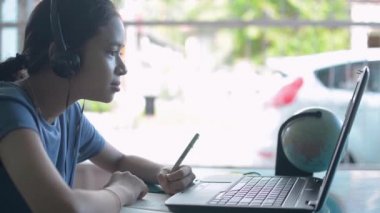 Kulaklık takan Asyalı sevimli liseli kız evde ders çalışırken bilgisayar kamerasıyla uzaktan öğretmenle konuşuyor. Modern internet teknolojisiyle uzaktan eğitim.