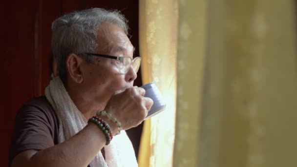 快乐的亚洲老年人白发苍苍 身穿休闲装 头戴眼镜 喝着热咖啡 在家中接受检疫时从窗口往外看 退休人员的生活方式 — 图库视频影像